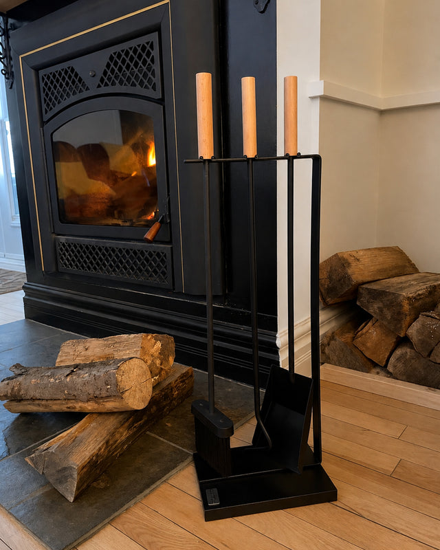 A black metal fireplace tool set with three light wood-handled tools stands on a wood floor in front of a black fireplace with a visible fire and stacked logs nearby.