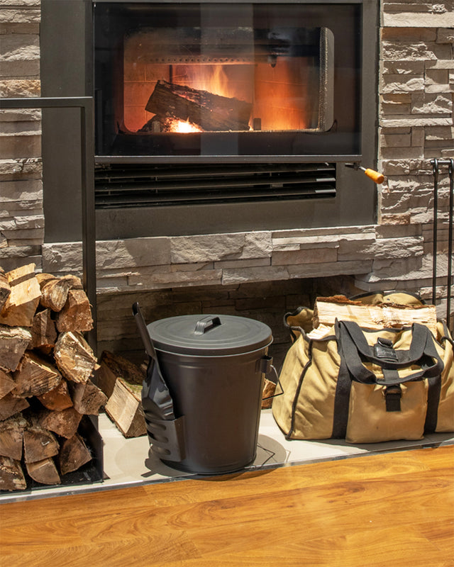 with-lid-hearth-the-rack-co.jpeg Black steel ash bucket with lid beside a wood-burning fireplace with log carrier bag and firewood rack