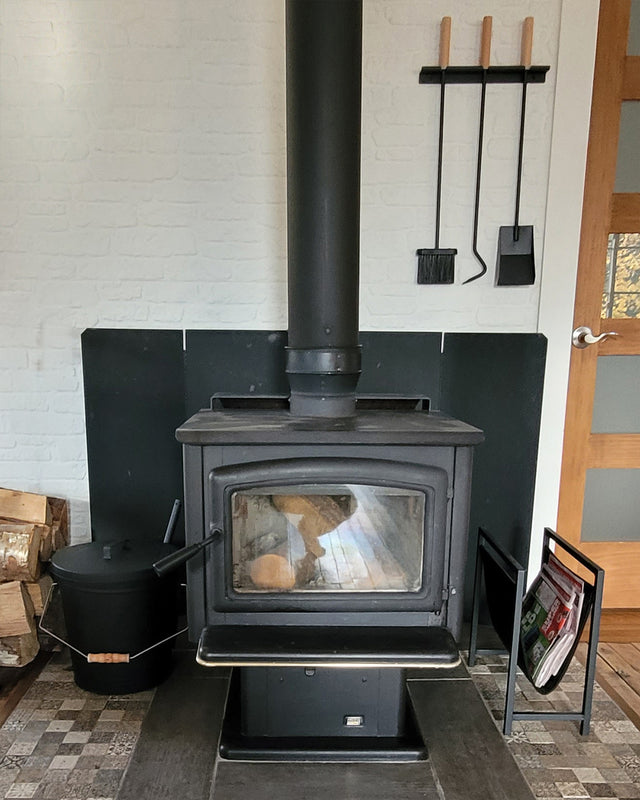 Fireplace ash bucket beside a wood-burning stove with firewood and fireplace tools