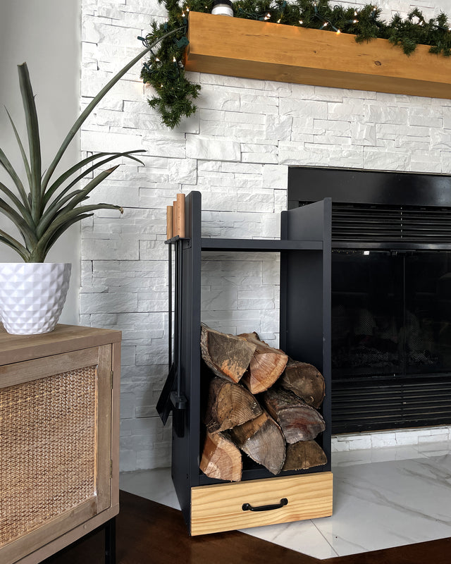 A black metal firewood storage rack with a light wood drawer base stands on a fireplace hearth beside a white brick modern farmhouse fireplace. The rack holds stacked firewood logs and has fireplace tools hanging on the left side.