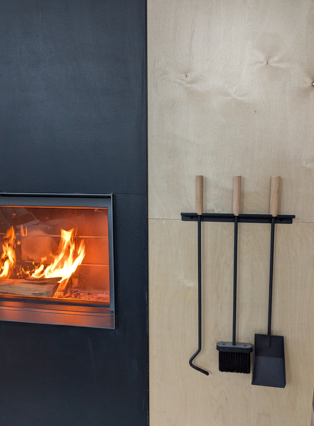 Wood-burning stove with fire burning inside, next to a set of wall mounted fireplace tools on a wood wall.