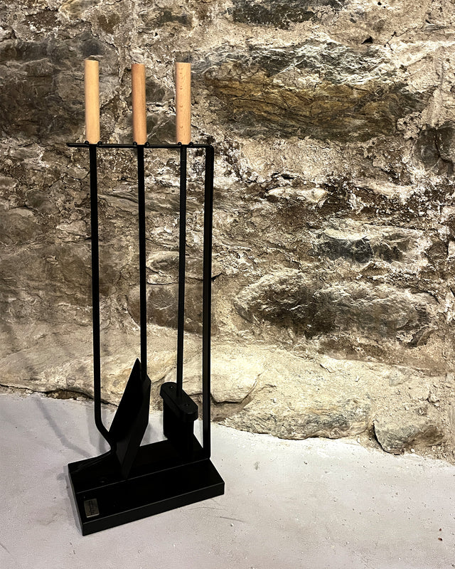 Modern 3-piece fireplace tool set with wooden handles displayed against a natural stone fireplace wall and light cement flooring.