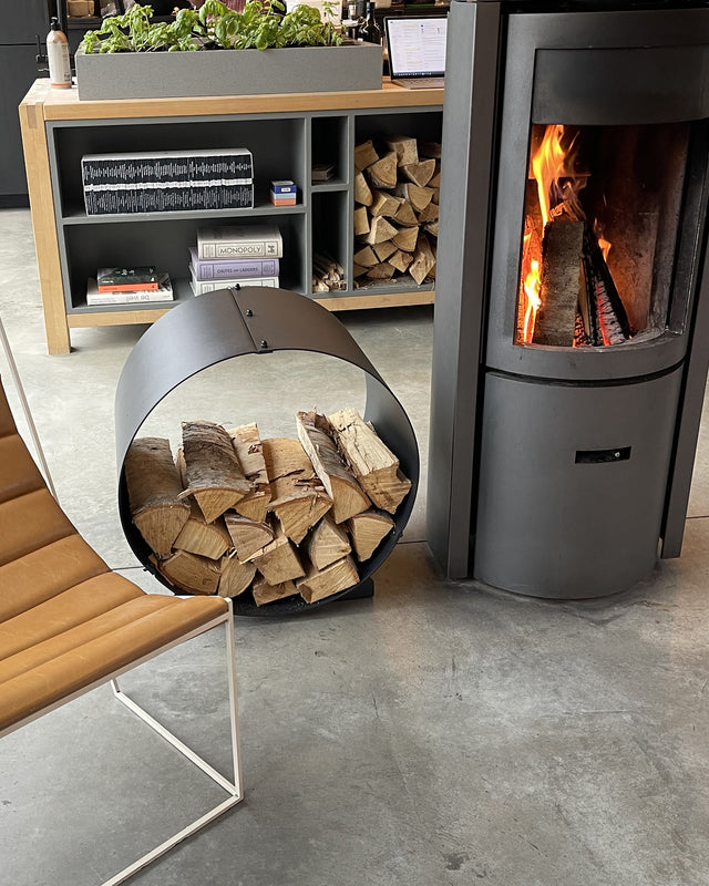 Black circular firewood rack storing logs beside a modern wood-burning stove in a modern cottage